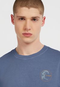 Blå bomulds T-shirt med rund hals og korte ærmer. Har et rundt logo med stiliseret tekst på venstre brystområde.