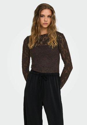 Jonge vrouw met golvend haar die een zwarte kanten top met lange mouwen en losse zwarte broek draagt tegen een effen lichte achtergrond.