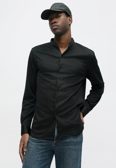 Nuori mies, joka pukee mustaa mandariinikauluksellista nappipaitaa, sinisiä farkkuja ja mustaa baseball-lakkia, seisoo yksinkertaista valkoista taustaa vasten.