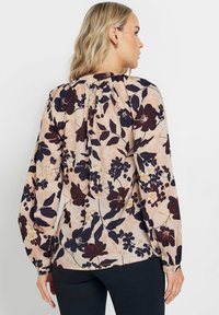 Blusa floreale in pesca chiaro con motivi viola scuro e gialli, caratterizzata da maniche lunghe a sbuffo, scollatura arricciata e polsini con bottoni.