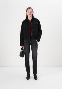 Juodas džinsinis švarkas su avikailio apykakle, dėvimas ant raudonos palaidinės, derinamas su juodomis siauromis kelnėmis ir juodais bateliais su smailiu noseliu. Laiko mažą juodą delninę.