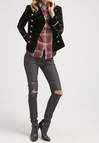 Giacca doppiopetto in velluto nero con bottoni dorati, camicia a quadri, jeans grigi strappati e stivali neri alla caviglia.