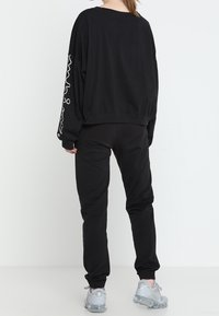 Schwarzer, übergroßer Sweatshirt mit langen Ärmeln, versehen mit einem weißen Grafikdesign. Kombiniert mit passenden schwarzen Jogginghosen und hellgrauen Sneakers.