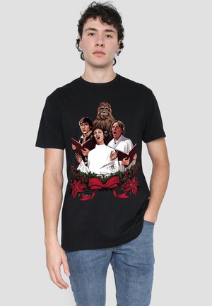 Black t-shirt featuring a colorful graphic of four characters singing, framed by holiday greenery and red ribbons. Cotton fabric, relaxed fit.