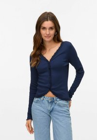 Marinblå långärmad cardigan med V-ringning, spetsdetaljer och knappstängning, kombinerad med ljusblå avslappnade jeans. Slät textur.