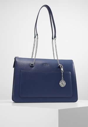 Sac à main DKNY bleu marine avec poche avant, bretelles chaîne argentée et pendentif rond argenté avec logo suspendu sur le côté droit.