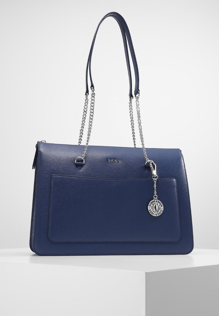 Sac à main DKNY bleu marine avec poche avant, bretelles chaîne argentée et pendentif rond argenté avec logo suspendu sur le côté droit.