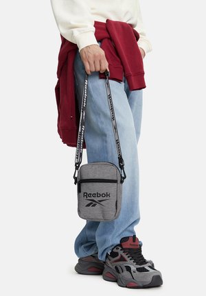 Osoba nosící světle modré džíny, šedé a červené tenisky Reebok, červenou mikinu svázanou v pase, držící šedou ramenní tašku Reebok.