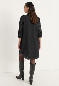 Schwarzes knielanges Kleid mit transparenter Überlagerung, strukturiertem Muster, ¾-Ärmeln und rundem Ausschnitt. Kombiniert mit knielangen schwarzen Lederstiefeln.