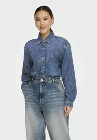 Camicia di jeans con bottoni e yoke in stile western, di colore azzurro chiaro e maniche lunghe. Indossata con jeans in denim sbiaditi a vita alta.