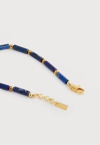 Collier avec perles cylindriques en pierre bleue, petites perles rondes en or, rallonge de chaîne en or et fermoir de homard en or sur fond blanc.