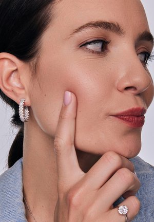 Srebrne kolczyki w kształcie kół z błyszczącymi detalami, gładka cera i naturalny makijaż, w połączeniu z dżinsową kurtką i pierścionkiem na lewej ręce.