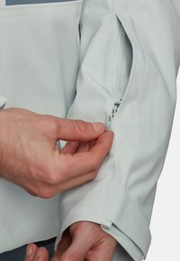 Veste imperméable gris clair avec une poche à zip sur la manche; poignets réglables. Le tissu présente une finition lisse et texturée.