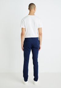 Un pantalon bleu marine en tissu lisse, au design slim-fit, avec deux poches arrière, associé à un t-shirt blanc uni et des chaussures blanches.