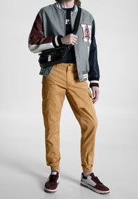 Varsityjack met grijze basis en bordeauxrode en marineblauwe mouwen, gecombineerd met mosterdkleurige cargobroek. Zwarte schoudertas, witte en rode sneakers.