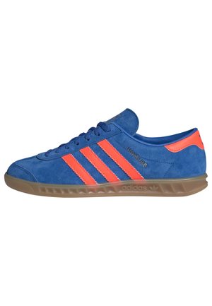 Blue suede sneaker with three bright orange stripes, rounded toe, gum sole, and branded accents on the side and heel.