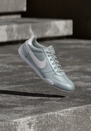 Világosszürke Nike sportcipő fehér pipa logóval, amely durva betonlépcsők és talaj felett lebeg.
