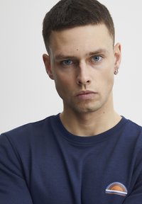 Tamsiai mėlynas džemperis su apvalia apykakle ir rausviais rankogaliais, su logotipo užrašu, kuriame yra oranžinės, rudos ir baltos spalvų akcentai. Lygus audinio paviršius.