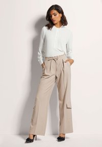 Witte lange mouwen blouse met knoopsluiting, gecombineerd met beige wijdvallende broek met zijzakken en een trekkoord in de taille. Zwarte hakken.