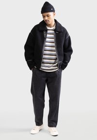 Black fleece jacket with a collar and zip, striped T-shirt, dark jeans, and white sneakers. Worn with a black knit beanie.
