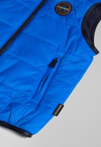 Blå puffer väst med hög krage, dragkedjeficka och logotyp patch. Har vadderad textur och slitstark yttertyg.