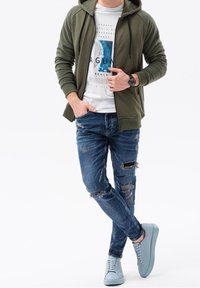 Felpa con cappuccio verde oliva con zip, t-shirt bianca con grafica, jeans blu strappati, scarpe da ginnastica azzurro chiaro, outfit casual su una persona.