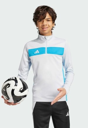 Camiseta deportiva blanca con cremallera y una franja horizontal azul, pantalones negros, sosteniendo un balón de fútbol negro y blanco, con el logo de Adidas.