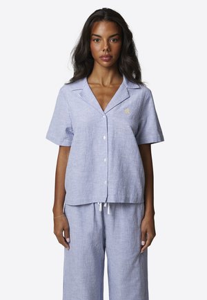 Kobieta ubrana w jasnoniebieską i białą koszulę na krótki rękaw z pionowymi paskami oraz dopasowane spodnie ze ściągaczem, zwrócona przodem.