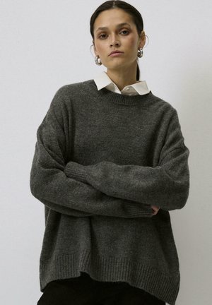 Mörkgrå oversized tröja i mjukt stickat material med rundad halsringning, ribbade ärmslut och fåll, lagd över en vit skjortkrage.