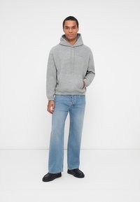 Sweat à capuche gris avec une poche avant, orné de broderies bleues. Associé à un jean bleu clair et des baskets noires, sur fond uni.