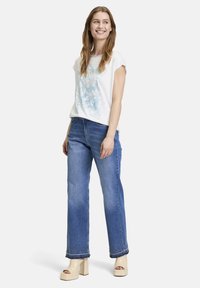White short-sleeve top with blue floral design and pattern, paired with blue straight-leg jeans featuring embellished hem and light platform heels.