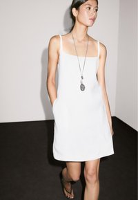 Weißes Leinenkleid mit schmalen Trägern, quadratischem Ausschnitt und seitlichen Taschen. Getragen mit einer langen silbernen Halskette und schwarzen Flip-Flops.