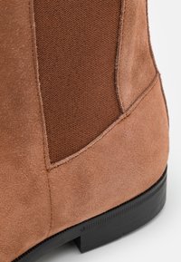 Bottine en daim marron avec un panneau en tissu texturé, une finition lisse, et une semelle en caoutchouc noire robuste avec un petit talon.