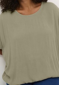 Mujer que lleva una blusa suelta de color verde oliva claro y de manga corta con cuello redondo, combinada con pantalones de color azul oscuro.