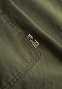 Olivno zelena tkanina z gladko teksturo in vidnim šivom; na spodnjem desnem kotu ima majhno etiketo z logotipom.