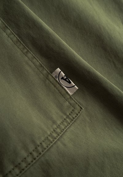 Tissu vert olive à texture lisse et coutures visibles ; comporte une petite étiquette avec un logo dans le coin inférieur droit.