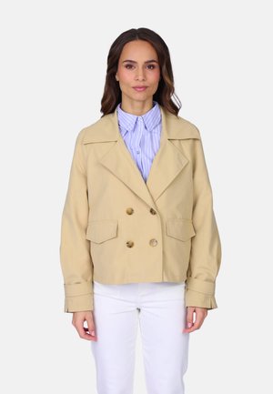 Femme portant une veste beige à double boutonnage sur une chemise rayée bleue et un pantalon blanc, debout devant un fond clair uni.