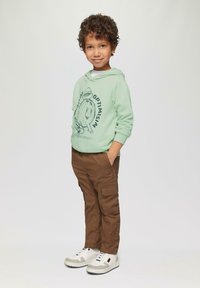 Mintgroene hoodie met een grafisch ontwerp, bruine cargobroek en witte sneakers met grijze accenten. Kind staat met handen in de zakken.