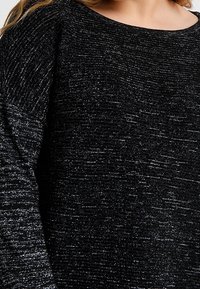 Černý dlouhý svetr s dlouhým rukávem se stříbrnými kovovými nitěmi, který nosí osoba se světlejší pokožkou a dlouhými vlasy.