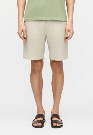 Muž v béžových kraťasoch, zelenej tričku a tmavohnedých sandáloch stojaci s uvoľnenými rukami po stranách na bielom pozadí.