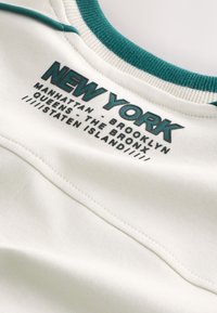 Tissu blanc avec un col accent turquoise. Le texte indique "NEW YORK" ainsi que les noms des arrondissements en impression en relief noire et turquoise.