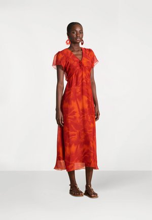 Femme portant une robe midi rouge-orange à imprimé tropical avec manches volantes, boucles d'oreilles rouges et sandales marron à brides, debout devant un fond uni.