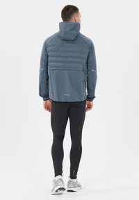 Mand iført en blå hættetrøje, sorte leggings, hvide sokker og hvide løbesko, som står og vender væk på en hvid baggrund.