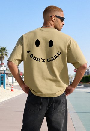 Mann trägt Sonnenbrille und beige T-Shirt mit schwarzem Smiley-Gesicht und "I DON'T CARE" Text auf dem Rücken, steht draußen in der Nähe von Palmen.