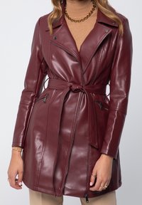 Donna che indossa una giacca di pelle bordeaux con cintura e tasche con zip sopra un dolcevita beige e pantaloni beige.