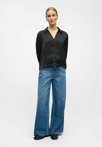 Blusa de satén negra de mangas largas con escote en V y cierre de botones, combinada con jeans de denim azul de pierna ancha y zapatos negros.
