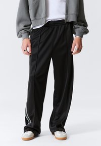 Schwarze Sporthose mit lockerem Schnitt, ausgestattet mit einem strukturierten Seitenstreifenmuster und einem elastischen Bund. Hergestellt aus glattem, leichtem Stoff.