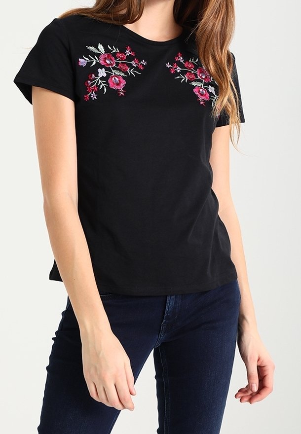 Femme portant un t-shirt noir à manches courtes avec une broderie florale rose et rouge sur le haut de la poitrine, associé à un jean bleu foncé.