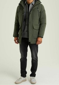 Giacca imbottita verde oliva con tasche con zip e bottoni a pressione, indossata sopra una camicia grigia con bottoni, abbinata a jeans neri e scarpe da ginnastica chiare.