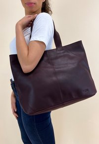 Dark burgundy leather tote bag with smooth texture, rectangular shape, and sturdy handles. Features subtle stitching and a logo on the side.